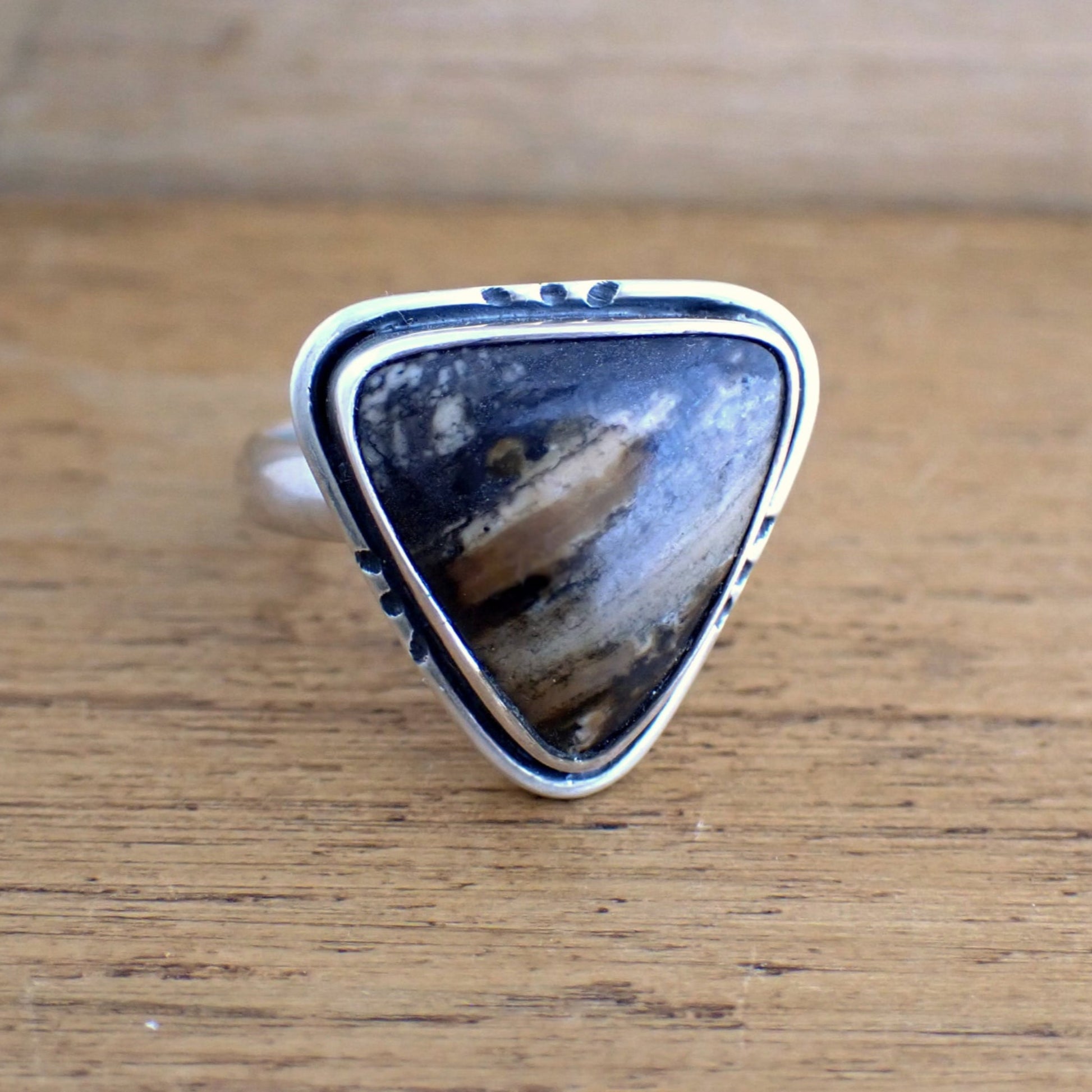 a ring on a raw wood surface - this features a large triangular stone with shades of brown, black, tan and cream running in streaks diagonally. The gemstone is set with the point downwards. Around the outside of the bezel of the stone is a groove oxidised black, and another slim silver frame of silver wire which has been engraved on all three sides with three notches (also blackened). The band of the ring is wide and rounded on the outside.