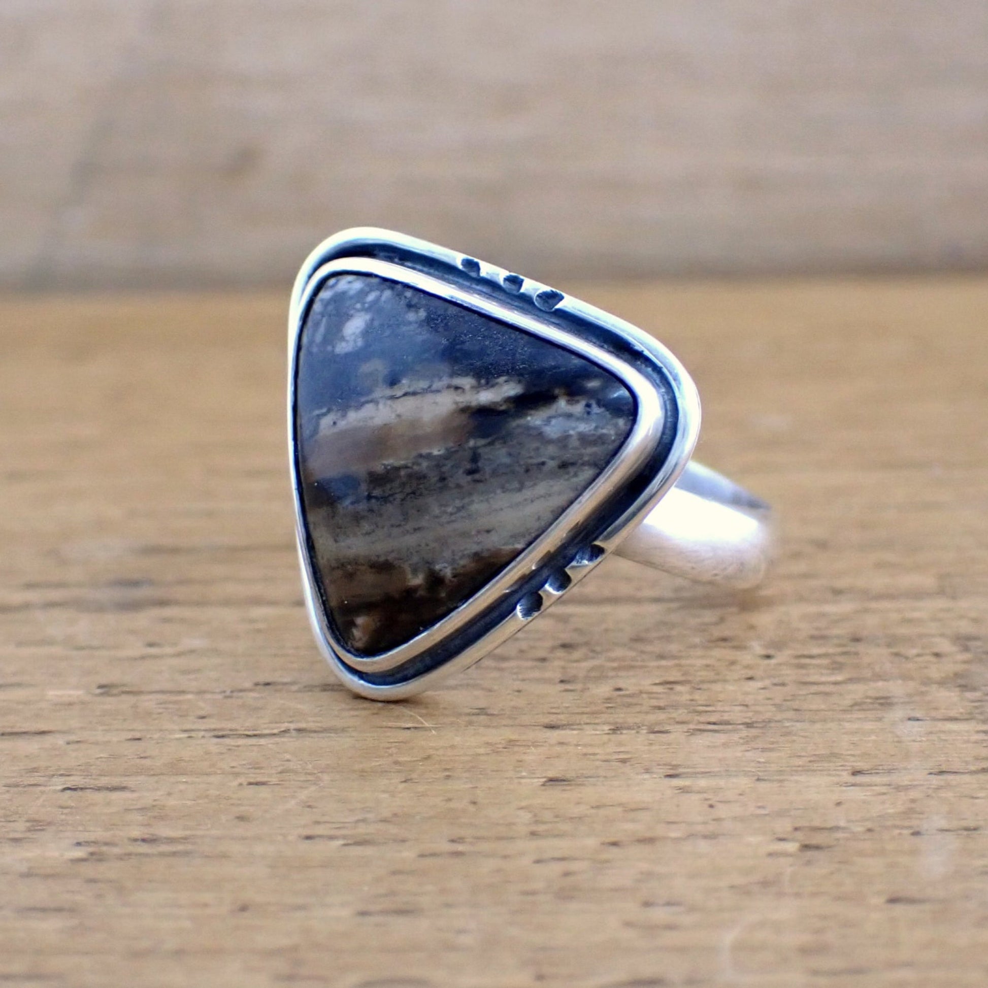 an alternative view of the Amaya ring on a raw wood surface - this photograph shows the detail of the gemstone more clearly - with patterns of black, brown, tan and cream in diagonal streaks and organic stipple like blotches