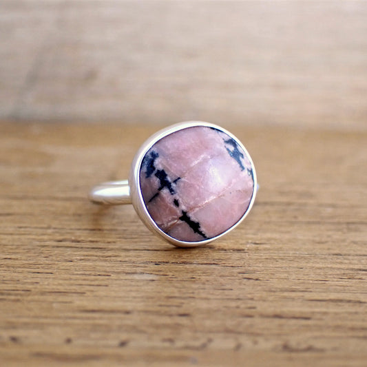 a small silver ring with a round soft pink and black opaque stone on a raw wood surface. This stone is predominantly pink with perhaps only 15 to 20% taken up with black manganese inclusions running diagonally across the ring. The band is simple, smooth and polished.