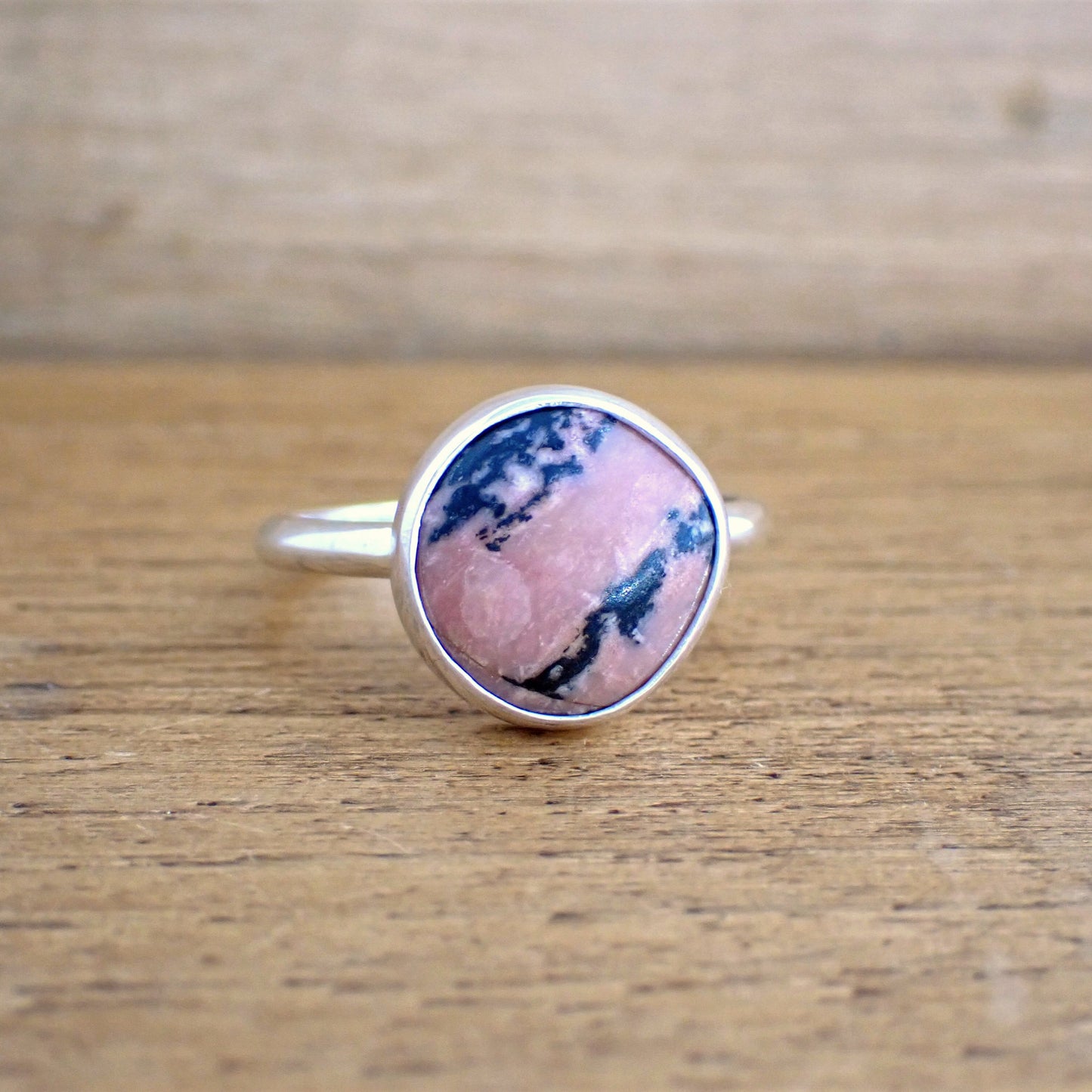 a small silver ring set with a pink and black opaque stone on a raw wood surface. The band of the ring is slim and smooth whilst the stone is set simply in a polished bezel. The gemstone is a soft mid pink colour, streaked with strong black inclusions running across it diagonally. There is also a patch of lighter pink on part of the face of the stone.