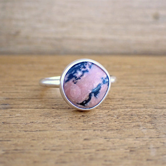 a small silver ring set with a pink and black opaque stone on a raw wood surface. The band of the ring is slim and smooth whilst the stone is set simply in a polished bezel. The gemstone is a soft mid pink colour, streaked with strong black inclusions running across it diagonally. There is also a patch of lighter pink on part of the face of the stone.