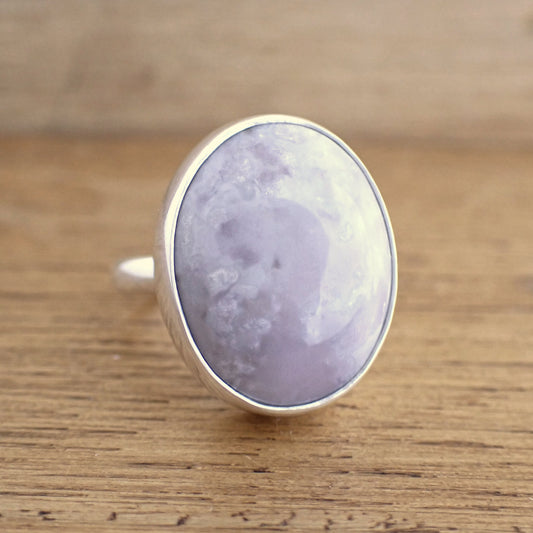A very large gemstone set ring on a raw wooden surface. This ring is set with a huge oval light purple stone which is highly polished and has patterns of lighter purple within it in plumey cloud shapes. The gem is set within a thick silver bezel on a plain band which are both also highly polished & shiny.