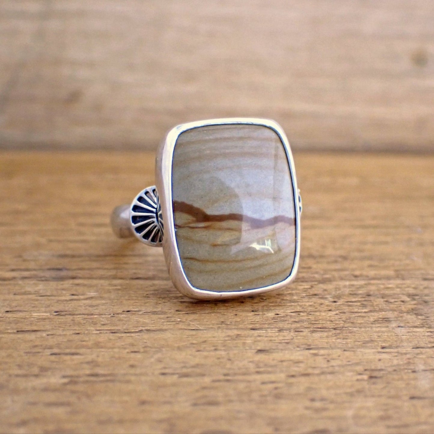A silver ring with a brown gemstone on a raw wood surface. The ring is set with a short rectangular stone in a shade of tan brown with darker streaks running through it giving the impression of hills / dunes and a sky. The stone is wrapped in a highly polished silver bezel and on the shoulders of the ring are stamp half-round domes resembling rays which radiate out from the stone. 