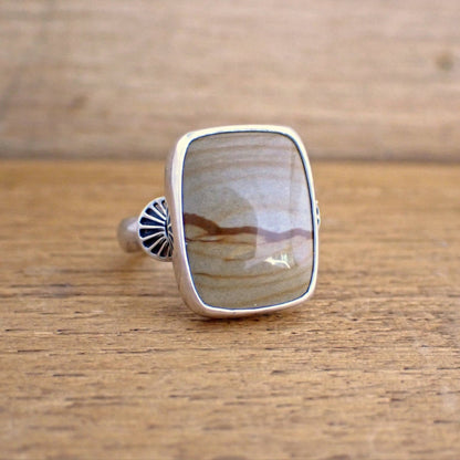 A silver ring with a brown gemstone on a raw wood surface. The ring is set with a short rectangular stone in a shade of tan brown with darker streaks running through it giving the impression of hills / dunes and a sky. The stone is wrapped in a highly polished silver bezel and on the shoulders of the ring are stamp half-round domes resembling rays which radiate out from the stone. 