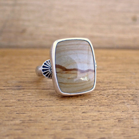 A silver ring with a brown gemstone on a raw wood surface. The ring is set with a short rectangular stone in a shade of tan brown with darker streaks running through it giving the impression of hills / dunes and a sky. The stone is wrapped in a highly polished silver bezel and on the shoulders of the ring are stamp half-round domes resembling rays which radiate out from the stone. 