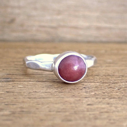 a small silver pink gemstone ring on a raw wooden surface. The ring band has been struck with wide hammered facets and is set with a small round deep pink gemstone which is opaque.