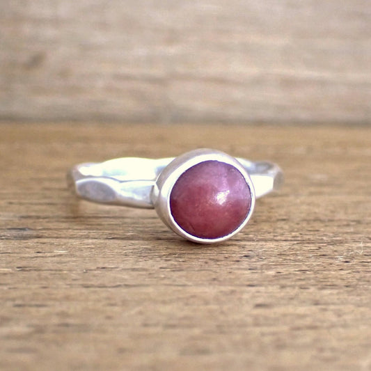 a small silver pink gemstone ring on a raw wooden surface. The ring band has been struck with wide hammered facets and is set with a small round deep pink gemstone which is opaque.