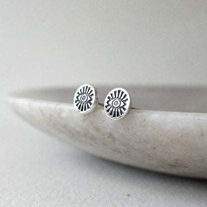 a pair of brighly polished silver stud earrings featuring a high detailed hand stamped eye - the eye design features long radiating eyelashes and has been darkened with patina - the earring is oval shaped - a quirky and fun design without being too out there!