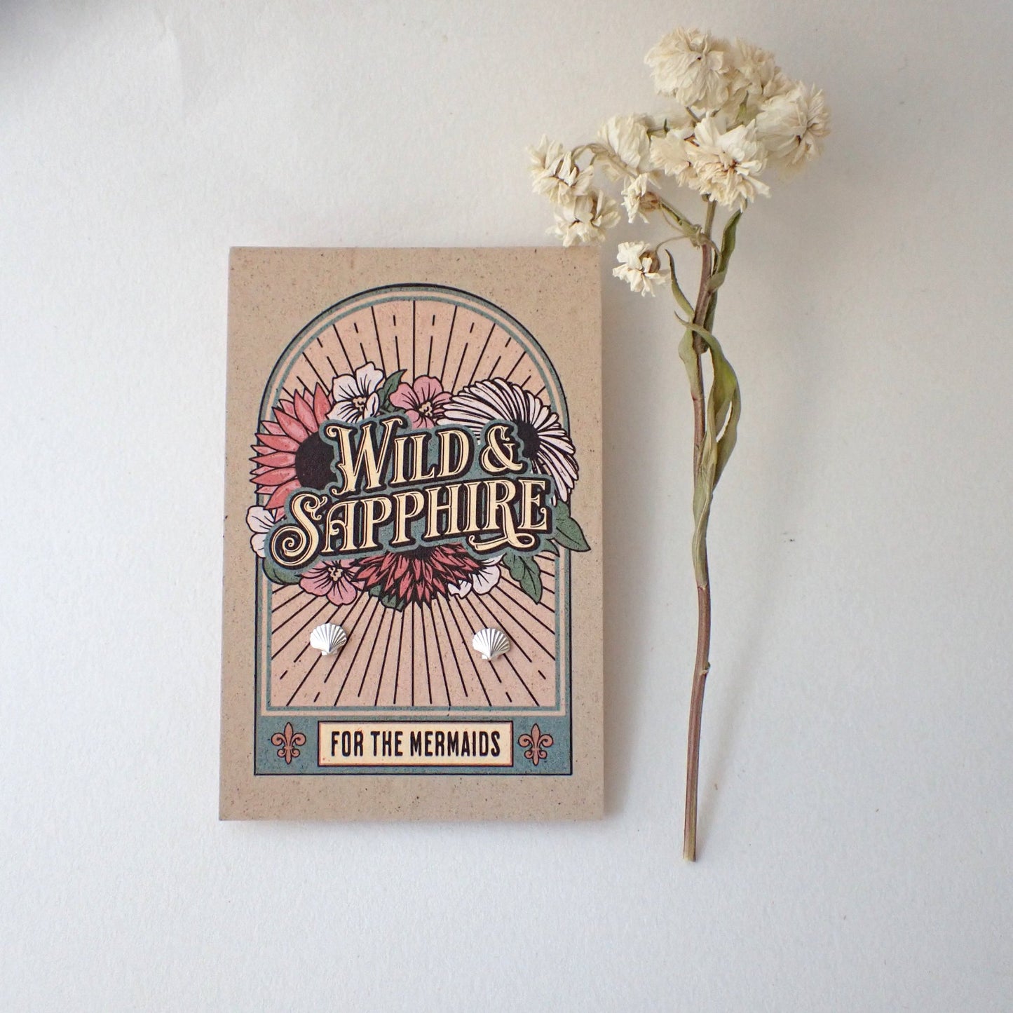 example of packaging - earrings mounted on festival inspired art card featuring 'Wild & Sapphire' surrounded by flowers and the caption "for the mermaids" - muted tones of blue, pink, peach and brown