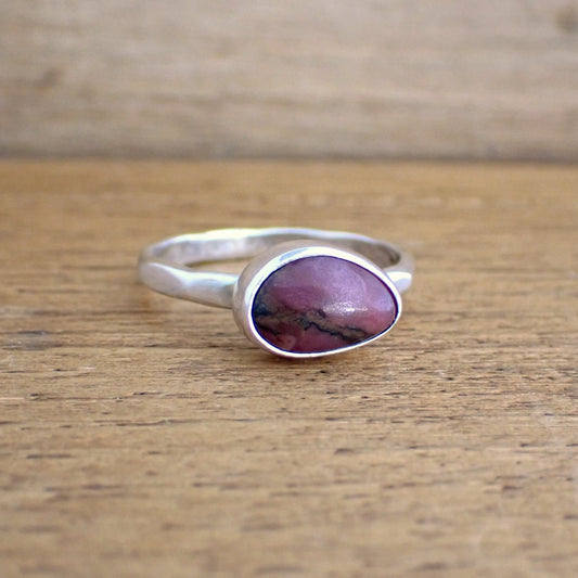 a small silver ring with a pink gemstone on a raw wooden surface. the ring band has wide hammered facets all the way round. The ring is set with a relatively small roughly pear shaped opaque gemstone in shades of dark pink with a black/brown streak running through it. The stone has been set sideways on the band which creates a chunkier look to the ring.